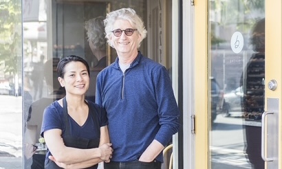 Woman and man, wearing blue, standing in front of b. Patisserie, outside.